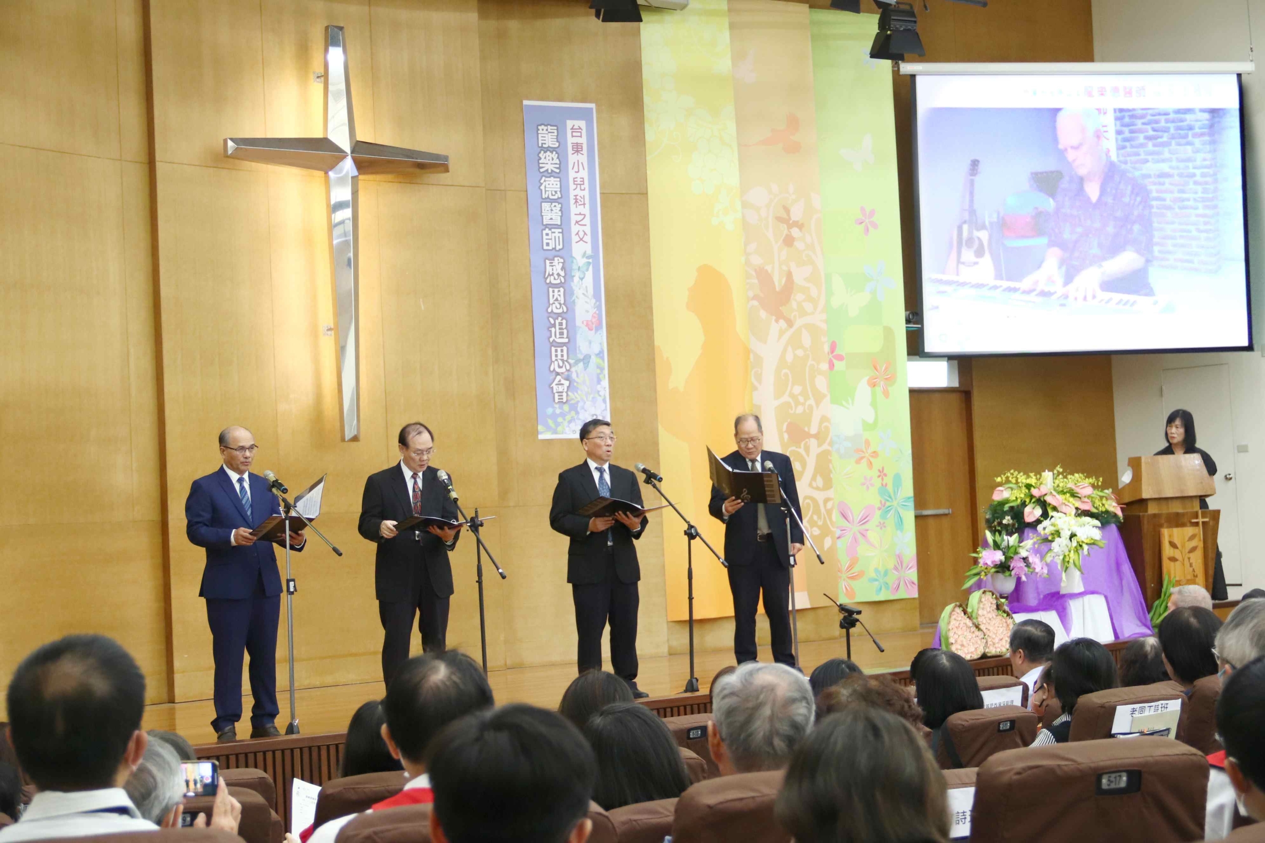 螢幕上播放龍醫師2016年彈奏「祢是我藏身處」的片段影片，緊接著由東基男聲四重唱上台獻唱「祢是我藏身處」等詩歌。（左起）李明福牧師、潘世隆牧師、東基院長馬堅毅、東基醫療財團法人執行長呂信雄。