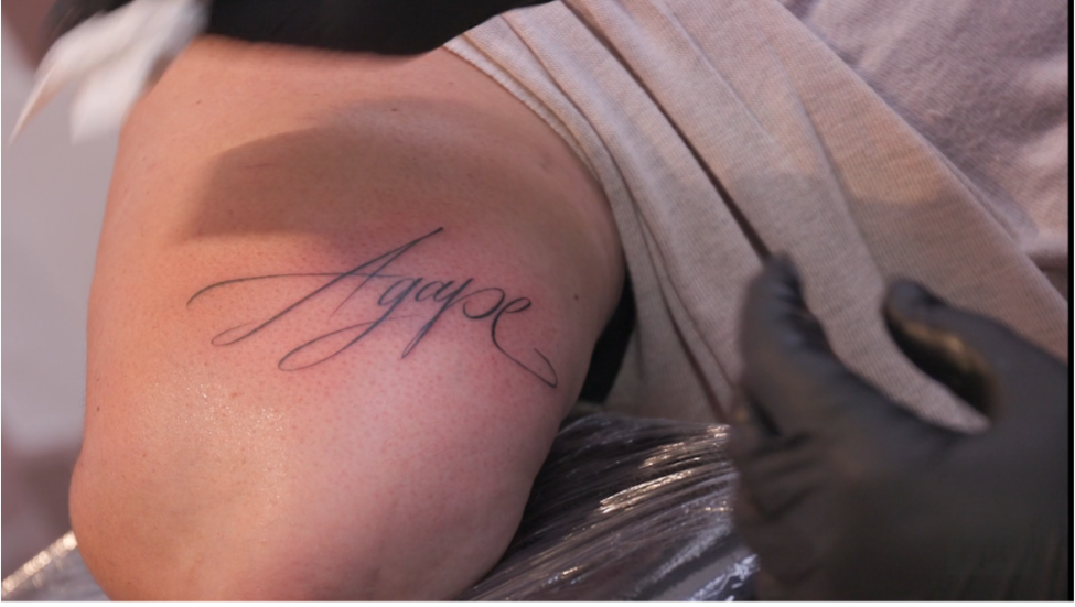 貝克斯為信徒安潔在手臂上紋「Agape」文字。（jesus.de影片截圖）