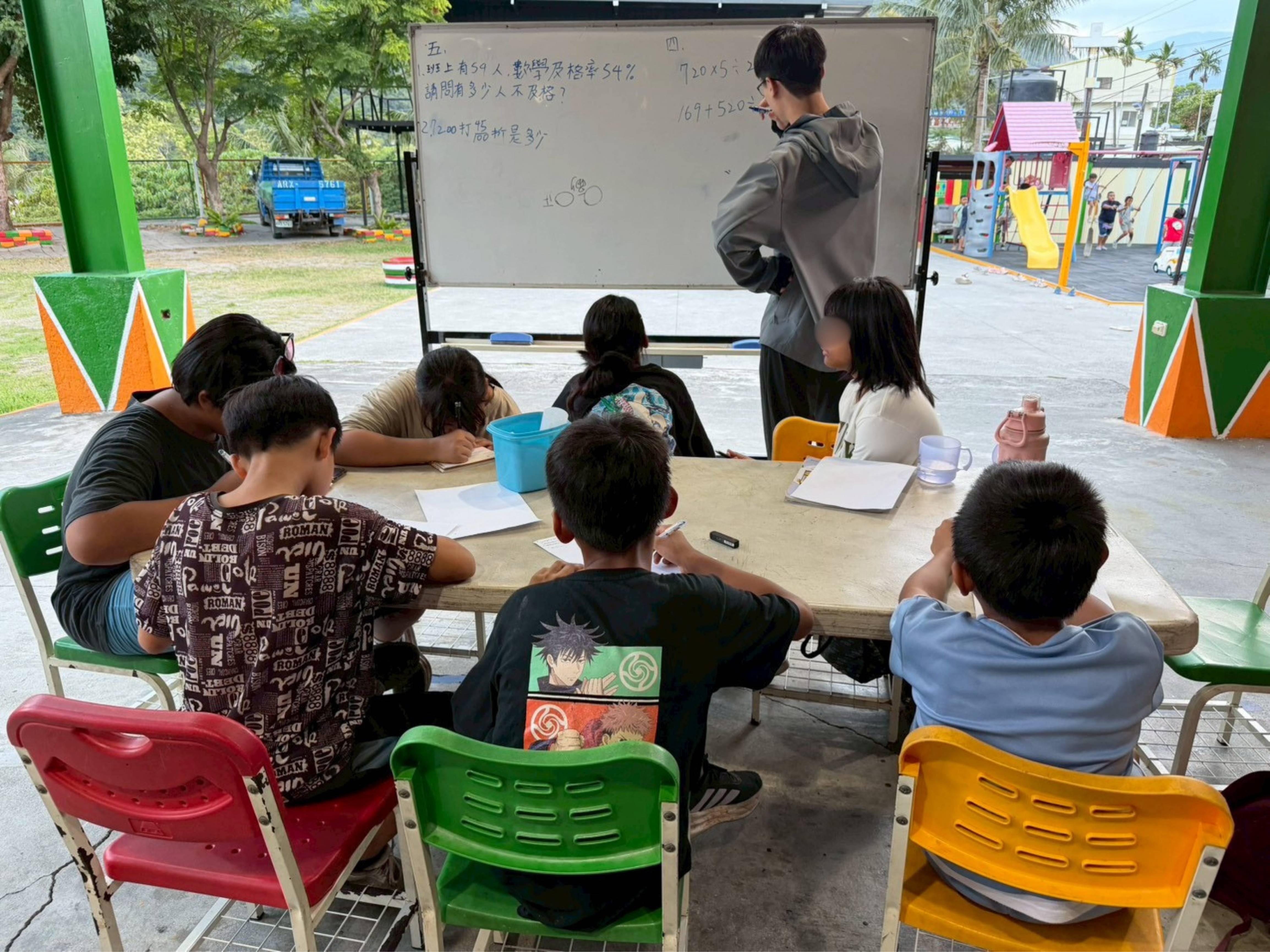 台東大學的吳沐恩同學在白板上寫下數學題，孩子們低頭專心解題，窗外籃球場的喧鬧完全不受影響。(圖/基督教救助協會提供)
